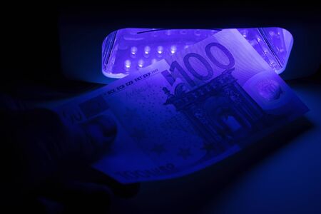 Verification of a banknote of 100 euros on a device for detecting fakes through ultraviolet light. Counterfeiting of banknotes, checking at the bank by the cashier, banking operator.の写真素材