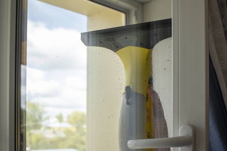 Wash the window at home with a battery wiper. The apparatus yellow collects drops of water and clean the window. Hands scrub the window. White window frame with a handle.の写真素材