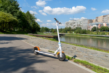 An electric white scooter stands on the bandwagon on the street. City park along the promenade....