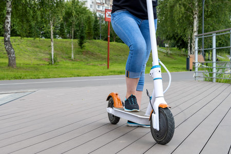 A girl in blue jeans rides a white electric scooter on the street. City park, summer sunny day,...