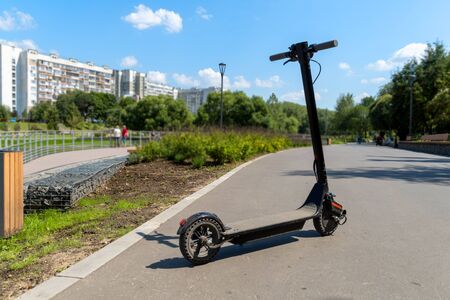 An electric black scooter stands on the bandwagon on the street. City park with wooden flooring along the promenade with railings. Sunny summer day. Modern city transport.の写真素材