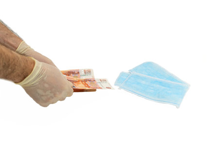 Two hands in white protective gloves buy a pack of blue medical masks for money with banknotes of...