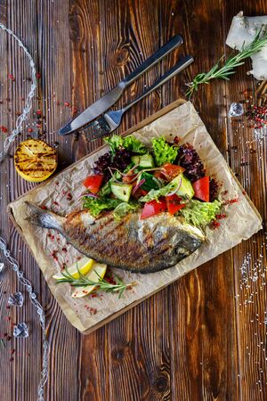 Grilled fish with vegetables and herbs on the kitchen board on a brown wooden backgroundの写真素材