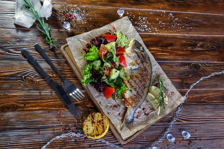 Grilled fish with vegetables and herbs on the kitchen board on a brown wooden backgroundの写真素材