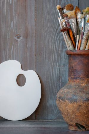 Brushes of the artist in an old clay pitcher and a white palette stand against the background of a wooden wall in the artist's studioの写真素材