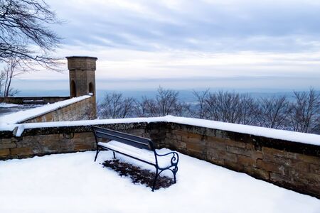 High in the mountains in the courtyard of Hohenzollern Castle stands a lonely bench covered with snow. On the ground is snow. In the background are mountains, blue sky and fluffy white clouds. Daytimeの写真素材