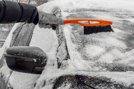 Man using Car Snow Brush to remove snow from the car windshield. Person in a black jacket and gray gloves. Close-up. Trees are covered with snow in the background. Winter, daytime.の写真素材