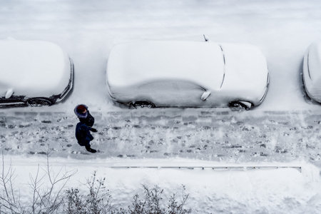 Parked cars and the road are covered with layer of snow. On the sidewalk are two people, a man and a...