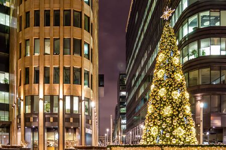 Christmas tree is decorated with lights, balls, toys and garlands, and star of gold color at the top. Facades of modern buildings in the background. Winter evening.の写真素材