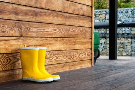 Blue and yellow rubber boots stand on the terrace. Adult and children's boots stand against the background of a wooden wall at home. Weather protection for your feetの写真素材