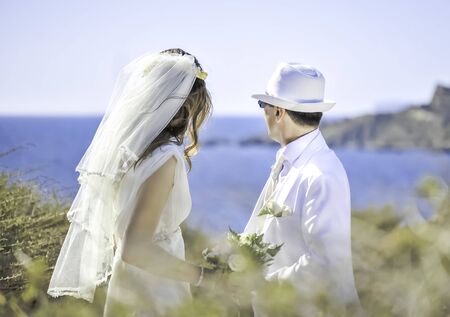 Summer wedding on Malta. Beautiful bride and groom on a cliff above the sea in a glamorous white wedding dress with scenic view to island, Malta. Happy woman, happy bride with scenic view in Malta. Daylight, outdoors, bright sun and blured blue sea at background.の写真素材
