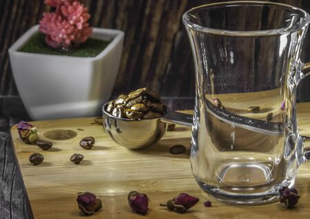 Measuring spoon for tea and coffee with coffee beans and dry tea leaves wth glass tea cup and copper cezve with orange wooden handle for Turkish coffee on a wooden plate. Jasmine, small roses, coffe beans and 2 pieces of sugar on the plate. Red plant at the background. Wooden background. Indoor. Mixed light.の写真素材
