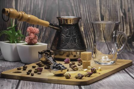 Measuring spoon for tea and coffee with coffee beans and dry tea leaves wth glass tea cup and copper cezve with orange wooden handle for Turkish coffee on a wooden plate. Jasmine, small roses, coffe beans and 2 pieces of sugar on the plate. Red and green plants at the background. Wooden background. Indoor. Mixed light.の写真素材