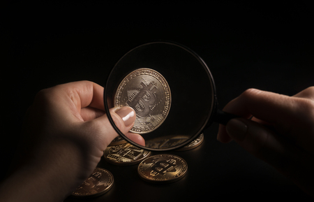 Unrecognizable person holding magnifying glass and looking through it on new virtual money golden bitcoin placed on a black background.の写真素材