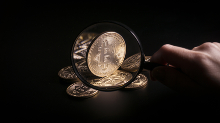 Unrecognizable person holding magnifying glass and looking through it on new virtual money golden bitcoin placed on a black background.の写真素材