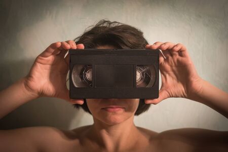 A young woman holding a video cassette in the face, like eyes. Grey backgroundの写真素材