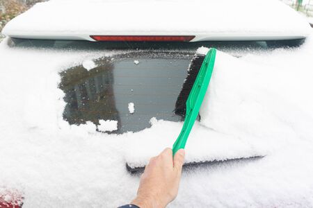 Man cleaning car from snow with brush. Snow-covered car windshield. parked car covered with snow during snowing in winter timeの写真素材