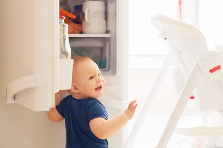one-year-old cute smiling blonde babe toddler boy opens the fridge while playing and looks what to eat.の写真素材