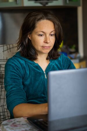 young woman in casual homewear working on laptop computer while sitting at the kitchenの写真素材