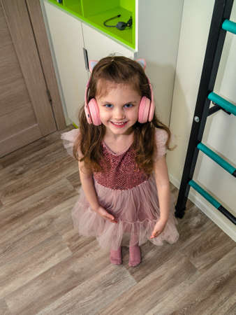 Little girl in a pink dress listening to wireless headphones at home.の写真素材