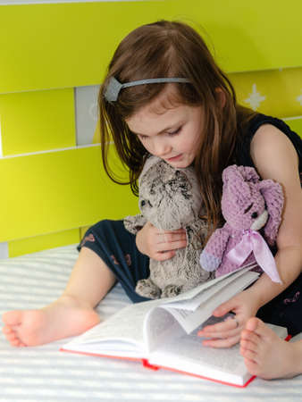A little girl is reading a book of fairy tales to her toys in her room on the bed.の写真素材