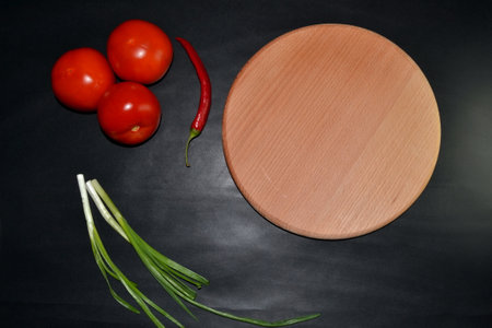 Green onions, red tomatoes, red hot pepper, round wooden board on dark background. Top view. Space for textの写真素材