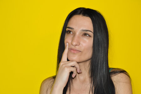 Young beautiful woman standing over isolated yellow background looking side confident, thinking with finger on a faceの写真素材