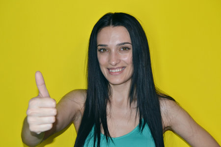 Happy brunette woman showing thumb up and looking at the camera over yellow background