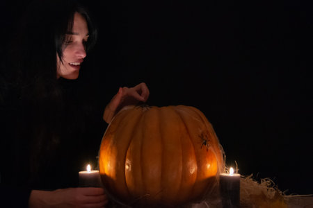 Dark hair pretty woman with big orange pumpkin on black background. The light of the candles illuminates her face. Conceptual photo of Halloween. Low key. Space for textの写真素材