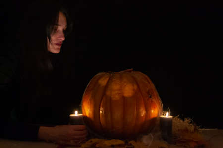 Dark hair pretty woman with big orange pumpkin on black background. The light of the candles illuminates her face. Conceptual photo of Halloween. Low key. Space for textの写真素材
