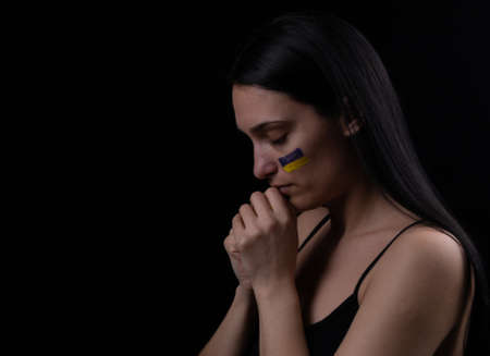 Portrait of ukrainian woman with blue and yellow ukrainian flag on her cheek praying for Ukraine. The symbol of the inviolability of the Ukrainian people. dark background. Space for textの写真素材