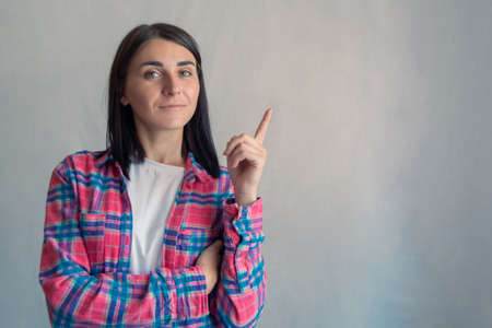Young attractive confident woman in casual shirt showing pointing finger at something or copy space empty text offer area. gray color background. Portrait of brunette hair pretty girlの写真素材