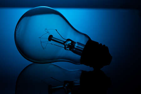 An incandescent light bulb lies on reflective glass, against a blue background, in a dark key.の写真素材