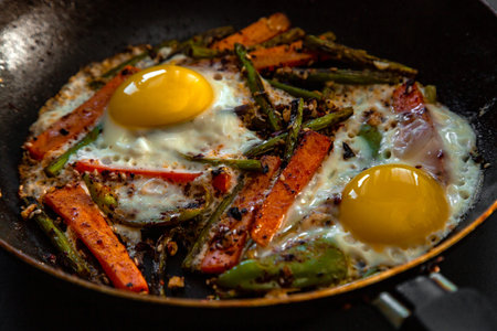 Very coarsely two fried eggs in a pan with large slices of carrots, bell peppers and asparagus with...