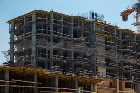 Two construction workers in helmets stand at the edge of a house under construction, against the...
