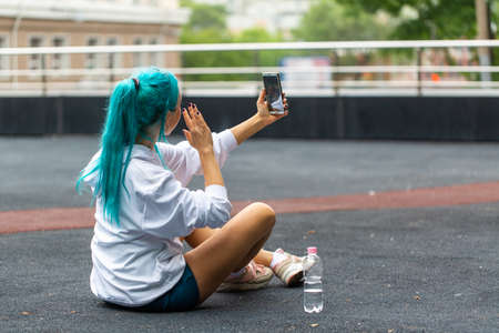A girl with blue hair talks to her subscribers through her phone online. Sitting on the ground in shorts.の写真素材