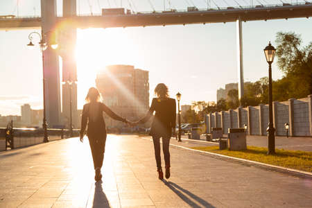 Two girls with blond hair walk along the boardwalk holding hands in the sunlight. Representatives of the LGBT community.の写真素材