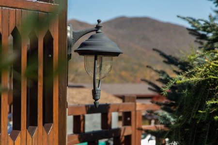An antique lantern with an LED lamp hangs on a wooden fence in front of a fir tree, in front of the mountains.の写真素材