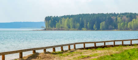 Walking path with a wooden fence along the shore of a large blue lake on a background of trees in summer,の写真素材