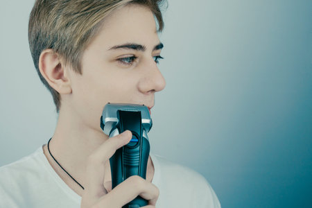 Portrait of a handsome smiling teenager in a white T-shirt shaves for the first time, shaves off his...