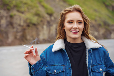 Smiling young woman in a denim jacket, with a cigarette in her hand near the road.