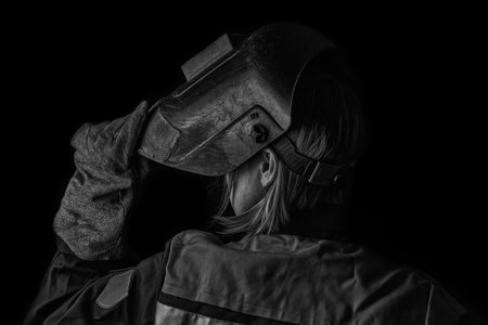 Welder man in the helmet and with welding terminals in the hand on the black wall background with copy space shows a thumbs up gesture.の写真素材