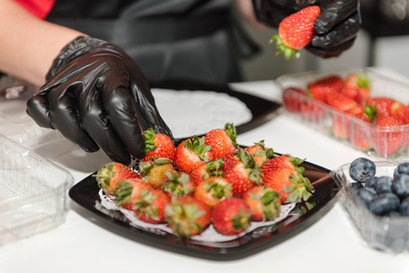 The girl puts strawberries on a plate. Juicy bright fresh strawberries on a plate. Cook puts the berry on a plate in a restaurant. The girl in gloves lays out the berries.の写真素材