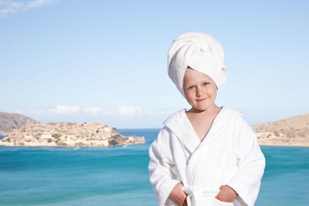 Portrait of happy smiling little girl with towel on the head in white bathrobe relaxing on terrace and looking at camera on the sea backgroundの写真素材