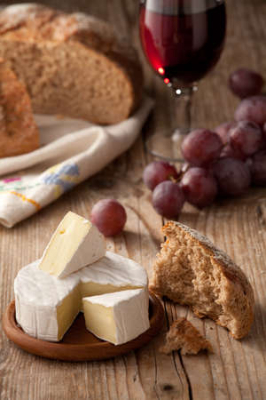 Piece and wheel of traditional Normandy soft cheese Camembert with country homemade bread, grapes and glass of red wine on wooden tableの写真素材