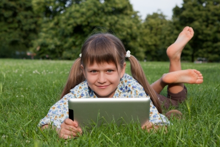 Cute smiling girl teenager holding tablet digital computer, looking at camera and relaxing on green grass in sunny parkの写真素材