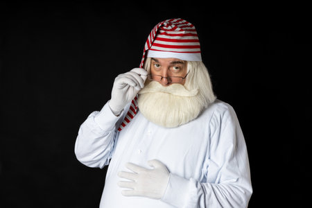 A bearded Santa man in a nightgown looks with an accent while holding his glasses. On a black...