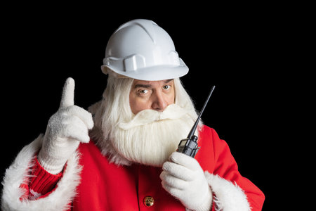 Santa Claus in construction helmet using walkie-talkie talking and pointing up on isolated black background, christmas industry concept.の写真素材