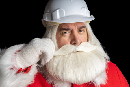 A stern man in a Santa suit with a gray beard and a helmet, adjusts his mustache with his hand and looks directly into the camera. Santa Claus industrial concept.の写真素材