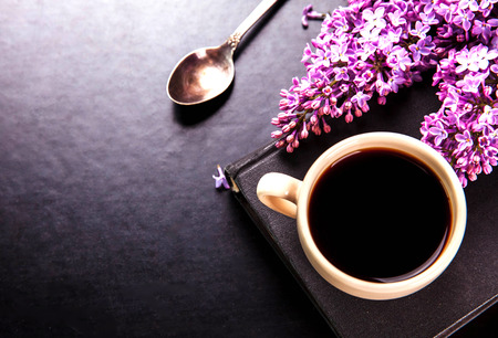 Black coffee in a cup, book, a spoon and fresh lilac flowers on black backgroundの写真素材